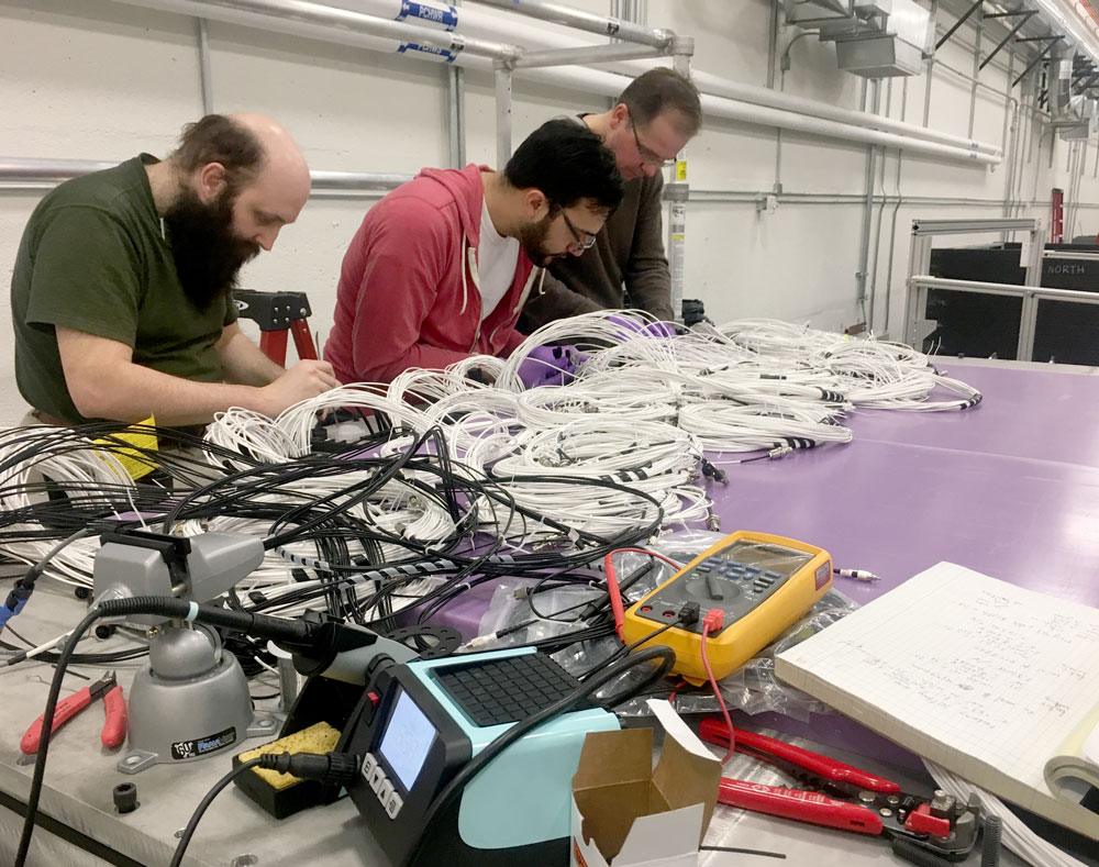 Three men work at a table covered in wiring, tools, and other components of the detector.