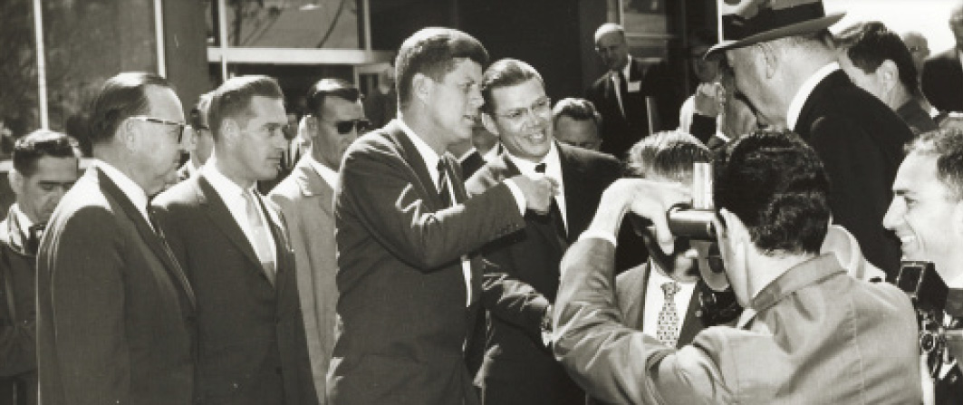 President John F. Kennedy stands before photographers alongside a half-dozen scientists.