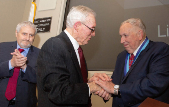 Two men shake hands at an awards ceremony while a third man looks on.