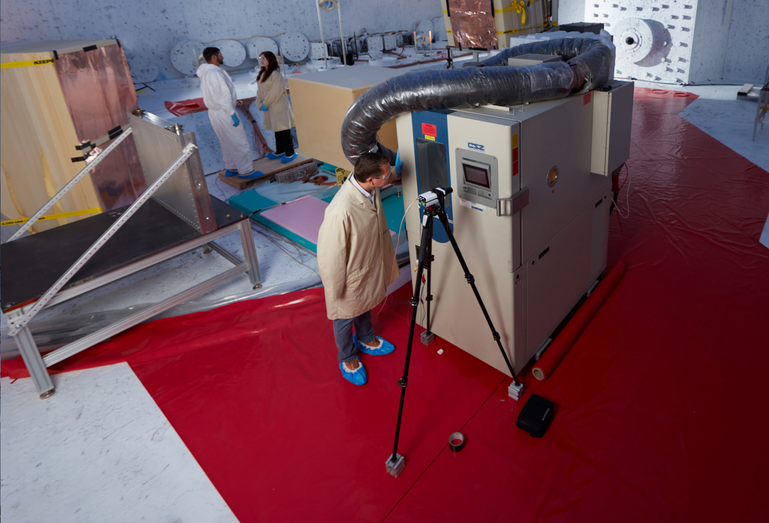 left: Scientists in a concrete room inspecting mirrors; right: A scientist inspects a large cubic machine in a concrete room.