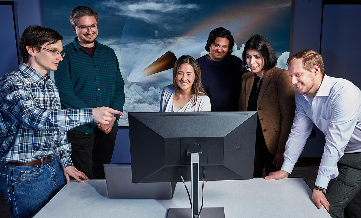 Photo of FPI teammates huddled amiably around a computer screen. Image on the wall behind them represents reentry vehicle in flight through clouds.