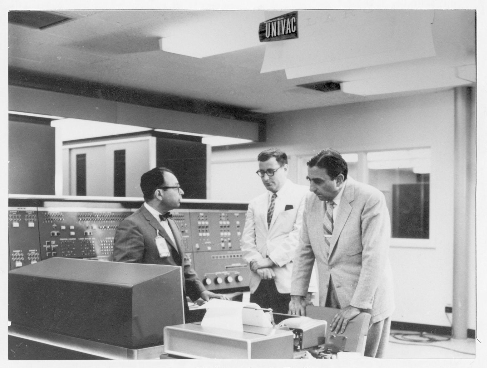Black and white archival photo of the three men standing in discussion in front of a computer control panel with knobs and switches.